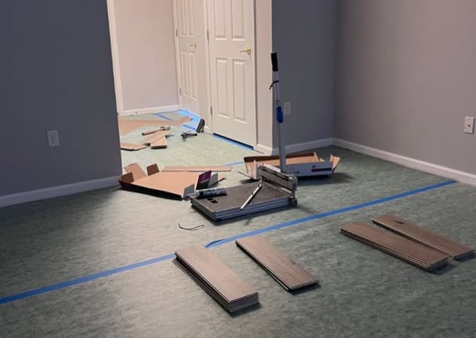Room under renovation with blue tape marking floor sections, tools, and laminate flooring samples laid out for installation.